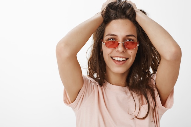 Freches attraktives Mädchen mit Sonnenbrille, die gegen die weiße Wand aufwirft