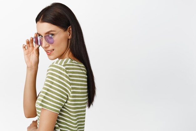 Freches brünettes Mädchen mit Sonnenbrille, das flirty in die Kamera blickt und im T-Shirt auf weißem Hintergrund steht