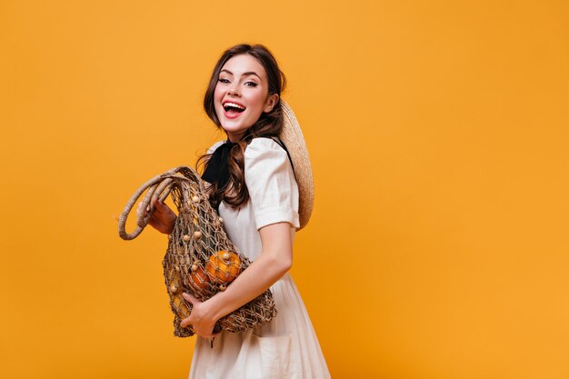 Freches dunkelhaariges Mädchen im weißen Kleid lacht und hält Öko-Tasche mit Produkten auf orange Hintergrund.