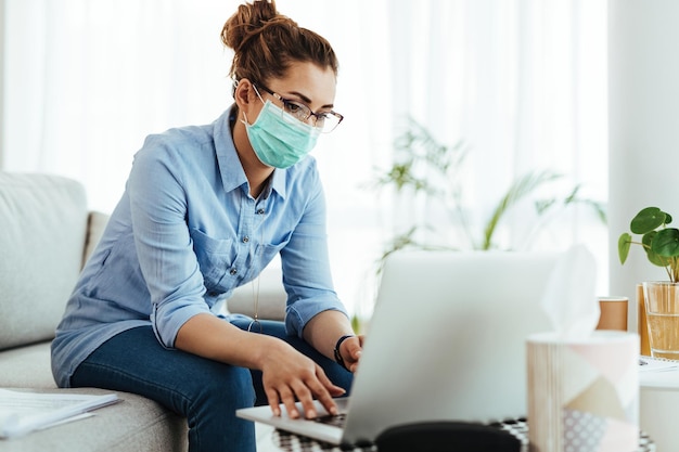Freiberuflerin mit Gesichtsmaske, während sie Laptop benutzt und während der Virusepidemie von zu Hause aus arbeitet