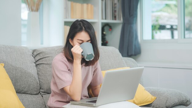 Freiberuflich tätige Asiatin des jungen Geschäfts, die an dem Laptop überprüft Social Media und trinkenden Kaffee beim Lügen auf dem Sofa arbeitet, wenn Sie sich im Wohnzimmer zu Hause entspannen. Lebensstilfrauen am Hauskonzept.