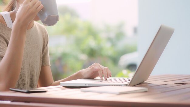 Freiberuflich tätige Asiatin, die zu Hause arbeitet, die Geschäftsfrau, die an Laptop arbeitet und den Kaffee sitzt auf Tabelle im Garten am Morgen trinkt.