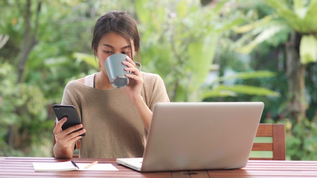 Freiberuflich tätige Asiatin, die zu Hause arbeitet, die Geschäftsfrau, die an Laptop arbeitet und den trinkenden Kaffee des Handys sitzt auf Tabelle im Garten am Morgen verwendet.