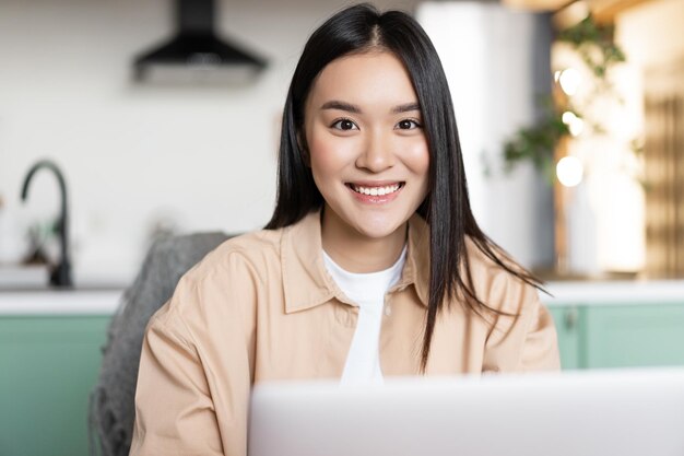 Freiberufliche asiatische mädchen, die zu hause sitzt und mit laptop arbeitet, lächelt in die kamera junge frau, die com ...