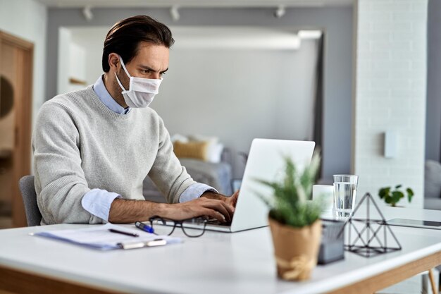 Freiberuflicher Mitarbeiter, der während der Coronavirus-Epidemie zu Hause mit Laptop arbeitet und eine schützende Gesichtsmaske trägt