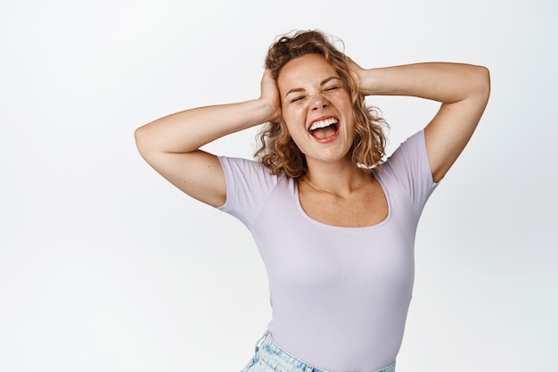 Freies und glückliches blondes Mädchen, das nachlässig schreit, die Hände auf dem Kopf hält, ihre Gefühle ausdrückt und im T-Shirt auf Weiß steht.