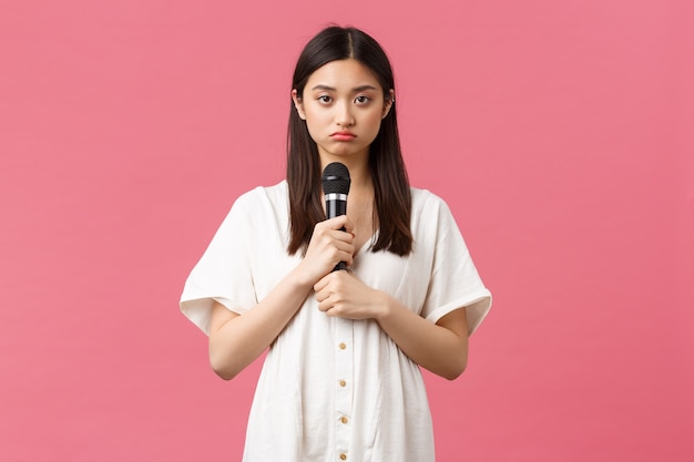 Freizeit, Emotionen und Lifestyle-Konzept. Düsteres und widerwilliges junges asiatisches mädchen, das mikrofon hält und traurige kamera schaut, nicht bereit ist, aufzutreten, stimmungsvoller rosa hintergrund steht.