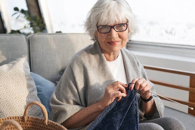 Freizeit, Hobby, Entspannung, Alter und handwerkliches Konzept. Fröhliche charmante Frau mittleren Alters im Ruhestand zu Hause entspannen, warmen Pullover zum Verkauf stricken, stilvolle Brille tragen und lächeln