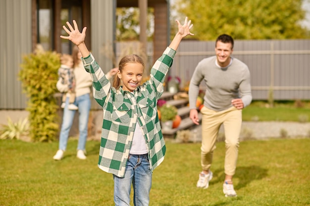 Freizeit. Lächelndes Mädchen im schulpflichtigen Alter in kariertem Hemd und Jeans, das die Hände hebt und in die Kamera schaut, Vater rennt und Mutter steht mit Kind hinten im Hof an einem sonnigen Tag