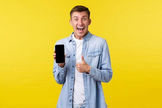 Freizeit-, Technologie- und Anwendungswerbekonzept. Aufgeregter, glücklicher und zufriedener blonder Kerl, der Daumen hoch zeigt, um eine super coole neue App zu empfehlen, die Online-Rabatte auf dem mobilen Bildschirm anzeigt.