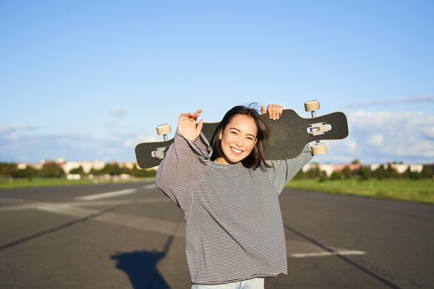 Kostenloses Foto freizeit und menschen glückliche asiatische frau, die mit longboard auf einer leeren straße in der landschaft steht