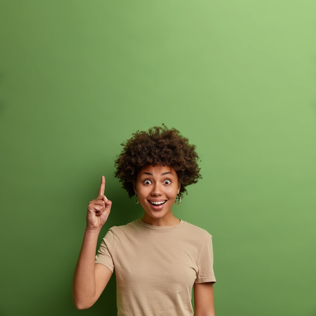 Freudig überrascht lockige erwachsene Frau zeigt auf leere Kopie Platz für kommerziellen Text, demonstriert Ideenpräsentation, tolle Promo nach oben, gekleidet in lässiges beiges T-Shirt, grüne Wand