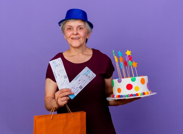 Freudige ältere Frau, die Partyhut trägt, hält Einkaufstasche des Papierkartenpapiers und Geburtstagstorte lokalisiert auf lila Wand mit Kopienraum