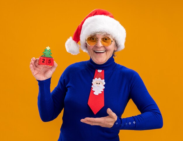 Freudige ältere Frau in Sonnenbrille mit Weihnachtsmütze und Weihnachtskrawatte, die auf Weihnachtsbaumverzierung lokalisiert auf orange Hintergrund mit Kopienraum hält und zeigt