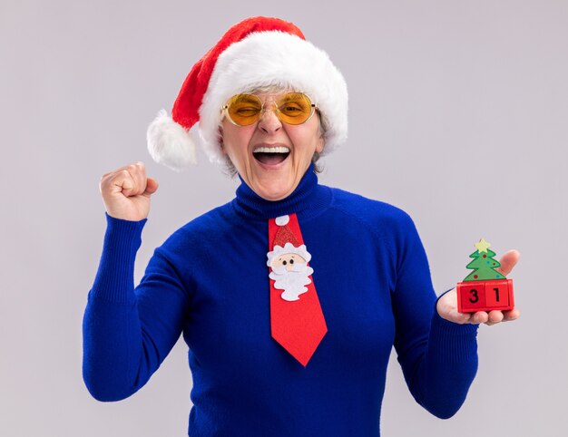 freudige ältere Frau in Sonnenbrille mit Weihnachtsmütze und Weihnachtskrawatte, die Weihnachtsbaumverzierung hält und Faust lokalisiert auf weißem Hintergrund mit Kopienraum hält