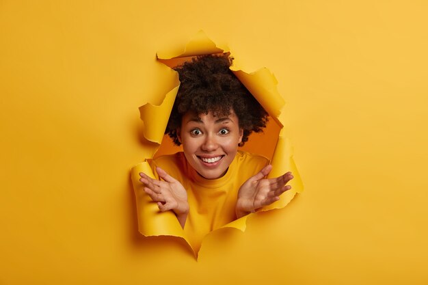 Freudige afroamerikanische Frau im gelben Pullover, spreizt Handflächen, zeigt weiße Zähne, hat Spaß drinnen, schaut durch zerrissenen Papierlochhintergrund