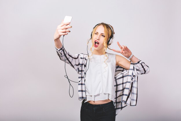 Freudige attraktive junge Frau, die selfie nimmt, Grimassen macht, Fingerfrieden singend zeigt. Fröhliches blondes Mädchen, das Spaß mit Smartphone hat. Helle Emotionen. Verrückte Partyzeit. Isoliert.