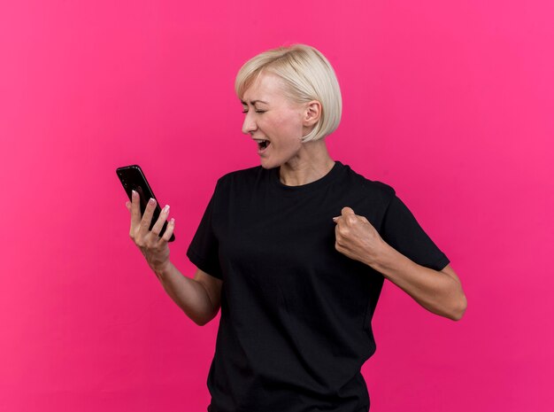 Freudige blonde slawische Frau mittleren Alters, die Handy hält, macht ja Geste mit geschlossenen Augen lokalisiert auf purpurrotem Hintergrund