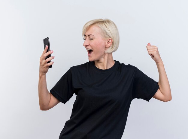 Freudige blonde slawische Frau mittleren Alters, die Handy hält, macht ja Geste mit geschlossenen Augen lokalisiert auf weißem Hintergrund