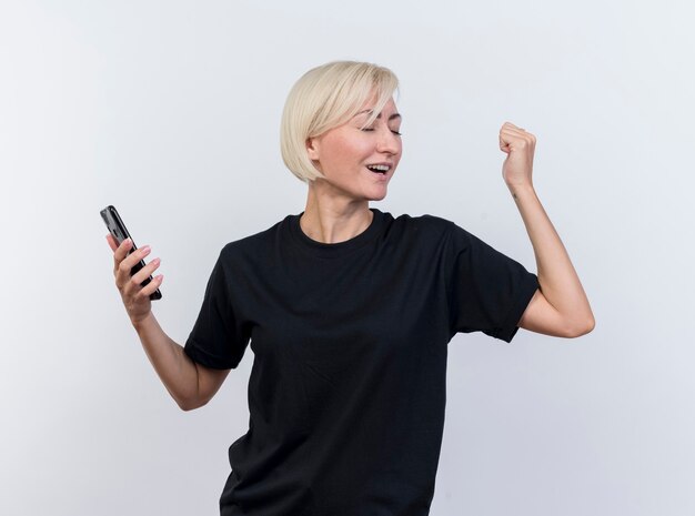 Freudige blonde slawische Frau mittleren Alters, die Handy hält, macht ja Geste mit geschlossenen Augen lokalisiert auf weißem Hintergrund