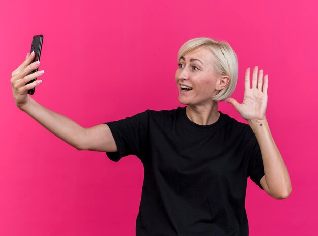 Freudige blonde slawische Frau mittleren Alters, die Handy hält und betrachtet, das hi Geste lokalisiert auf rosa Wand tut