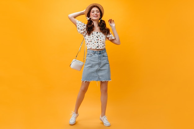 Freudige Dame, gekleidet in Jeansrock und verkürztes Oberteil, das mit weißer Tasche auf orange Hintergrund aufwirft.