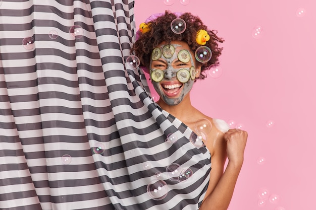 Kostenloses Foto freudige dunkelhäutige frau mit afro-haaren trägt tonmaske mit gurkenscheiben auf