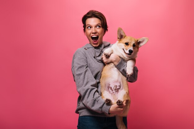 Freudige Frau im grauen Kapuzenpulli, der Corgi auf rosa Hintergrund hält. Emotionales jugendlich Mädchen in Jeans spielt mit Hund auf isoliert