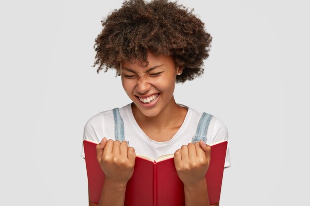 Freudige Frau lacht glücklich, während sie lustige Geschichte aus dem Buch liest, zeigt weiße Zähne, blinzelt Gesicht als Lächeln, gekleidet in lässigem Outfit, isoliert über weißer Wand. Menschen, Hobby und Lesekonzept