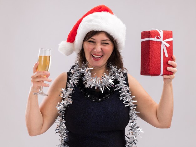 Freudige Frau mittleren Alters, die Weihnachtsmütze und Lametta-Girlande um den Hals hält, hält Weihnachtsgeschenkpaket und Glas Champagner, die Kamera betrachten, die lokalisiert auf weißem Hintergrund zwinkert
