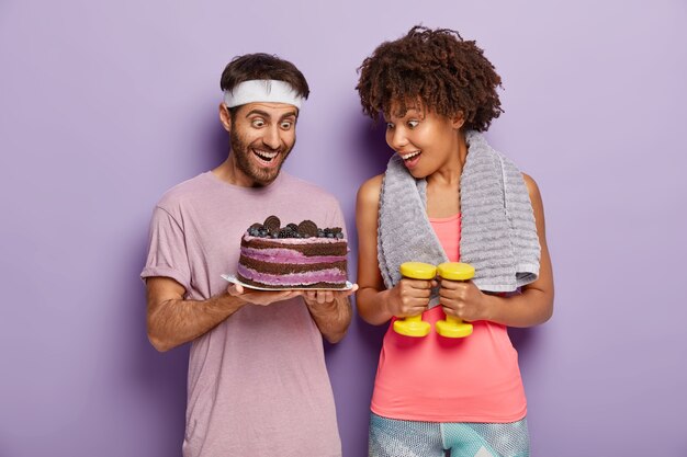 Freudige Frauen und Männer starren mit Glück und Versuchung auf köstlichen Kuchen, sind nach erschöpftem Training hungrig, vermeiden es, süße Desserts mit viel Kalorien zu essen, trainieren Sie mit Hanteln im Fitnessstudio