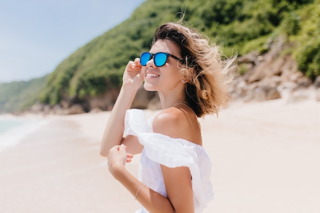 Freudige gebräunte frau mit hellbraunem haar, das sich beim aufstellen im resort umschaut. prächtige junge frau in der sonnenbrille lächelnd