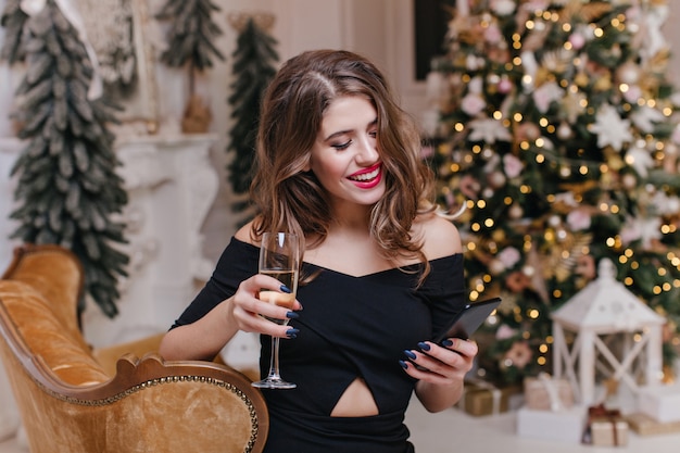 Freudige, glückliche junge Frau in teuren Kleidern trinkt leckeren Weißwein und liest an Weihnachten angenehme Grüße in ihrem Handy