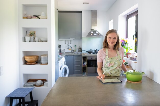 Freudige Hausfrau, die Notizen auf Block für Rezept schreibt, während sie in ihrer Küche kocht, Tablette nahe großem Topf auf Zähler verwendet, Kamera betrachtet. Vorderansicht. Kochen zu Hause und Online-Kochbuchkonzept