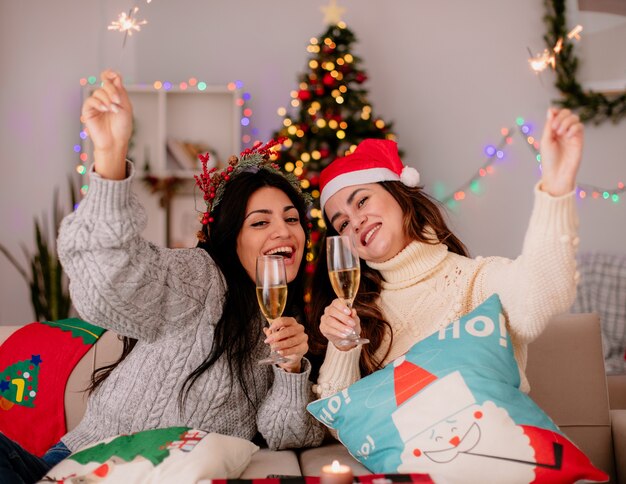freudige hübsche junge Mädchen halten Gläser Champagner und Wunderkerzen, die auf Sesseln sitzen und die Weihnachtszeit zu Hause genießen