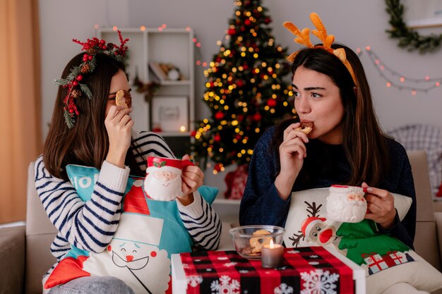 freudige hübsche junge Mädchen mit Stechpalmenkranz und Rentierstirnband halten Tassen und essen Kekse, die auf Sesseln sitzen und die Weihnachtszeit zu Hause genießen