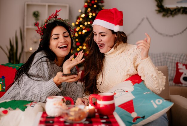 Freudige hübsche junge Mädchen mit Weihnachtsmütze schauen auf Telefon, das auf Sesseln sitzt und Weihnachtszeit zu Hause genießt
