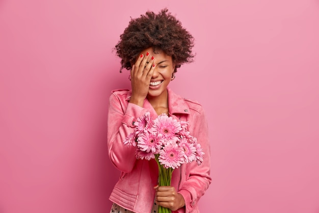 Kostenloses Foto freudige junge afroamerikanerin lacht glücklich, macht gesichtspalme, bekommt schönes geschenk wie blumen, hält schöne gerbera, drückt aufrichtige gefühle aus,