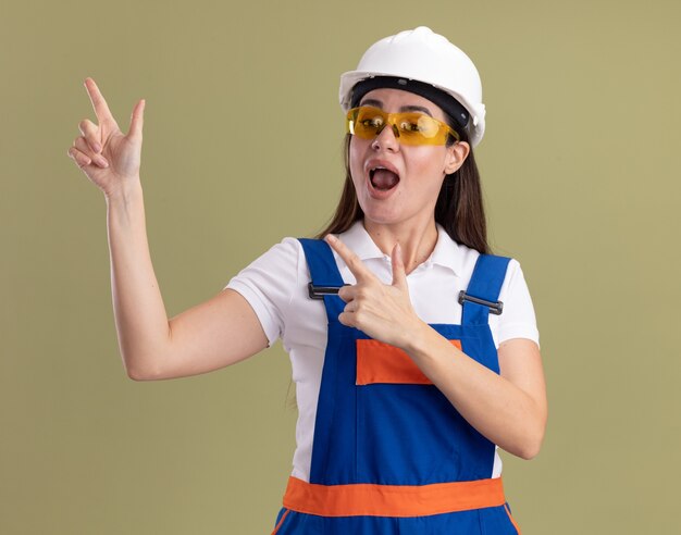 Freudige junge Baumeisterin in Uniform und Brille zeigt an der Seite lokalisiert auf olivgrüner Wand mit Kopienraum