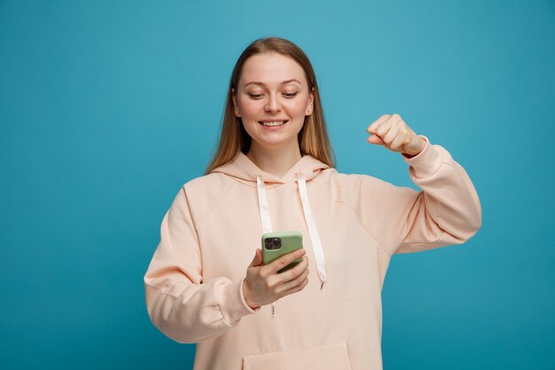 Freudige junge blonde Frau hält und schaut auf Handy, das ja Geste tut