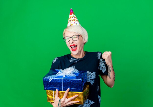 Freudige junge blonde Partyfrau, die Brille und Geburtstagskappe hält, die Geschenkboxen hält, die ja Geste lokalisiert auf grüner Wand mit Kopienraum tun