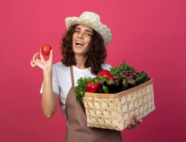 Freudige junge Gärtnerin in Uniform mit Gartenhut, die Gemüsekorb mit Tomate lokalisiert auf Rosa hält