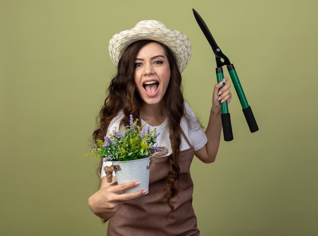 Freudige junge Gärtnerin in Uniform mit Gartenhut hält Blumentopf und Gartenschere isoliert auf olivgrüner Wand