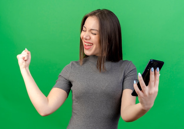 Freudige junge hübsche Frau, die Handy hält, das Seite betrachtet, die ja Geste mit geschlossenen Augen lokalisiert auf grünem Hintergrund tut