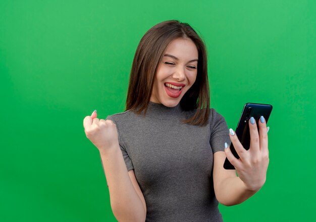 Freudige junge hübsche Frau, die Handy hält und betrachtet und Ja-Gesten-Telefon lokalisiert auf grünem Hintergrund mit Kopienraum tut