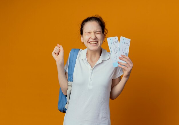 Freudige junge hübsche Studentin, die Rückentasche hält, die Flugtickets hält, die Faust mit geschlossenen Augen bündeln lokalisiert auf orange Hintergrund mit Kopienraum