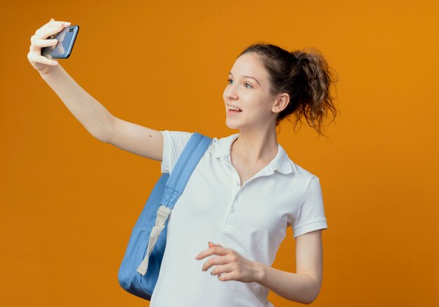 freudige junge hübsche Studentin, die Rückentasche trägt und Selfie nimmt, das Hand in Luft hält