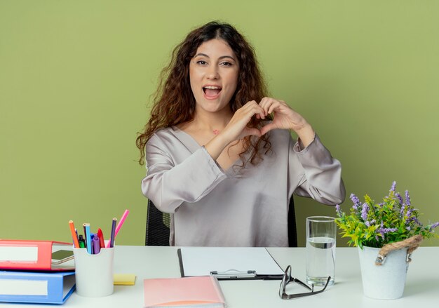 Freudige junge hübsche weibliche Büroangestellte, die am Schreibtisch mit Bürowerkzeugen sitzt, die Herzgeste lokalisiert auf olivgrünem Hintergrund zeigen