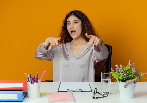 Freudige junge hübsche weibliche Büroangestellte, die am Schreibtisch mit Bürowerkzeugpunkten an der Kamera lokalisiert auf orange Hintergrund sitzt