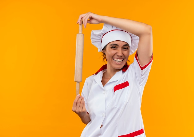 Freudige junge Köchin, die Kochuniform trägt, die Nudelholz auf isolierter gelber Wand mit Kopienraum hält